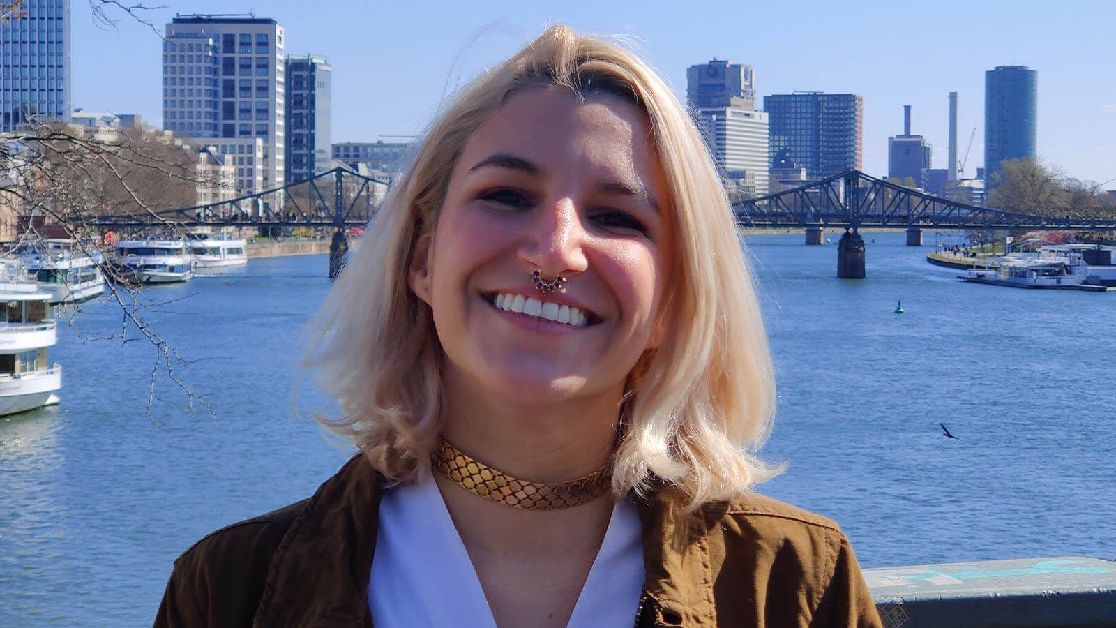 Grad Angela Decastro, a woman with shoulder-length blonde hair wearing a brown jacket and white top, smiles while standing on a bridge overlooking a river, in the distance behind her is a bridge and cityscape.