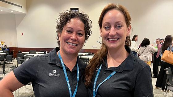 Michelle Moreno and Stacie Aldrich smiling and standing side by side wearing blue lanyards with conference badges and black Full Sail polo shirts.